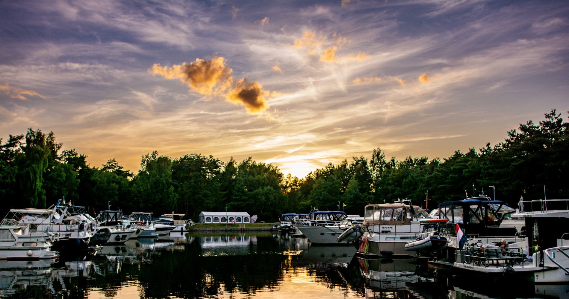 Haven met diverse boten aan weerszijden, omgeven door bomen, bij zonsondergang (vergroot foto)